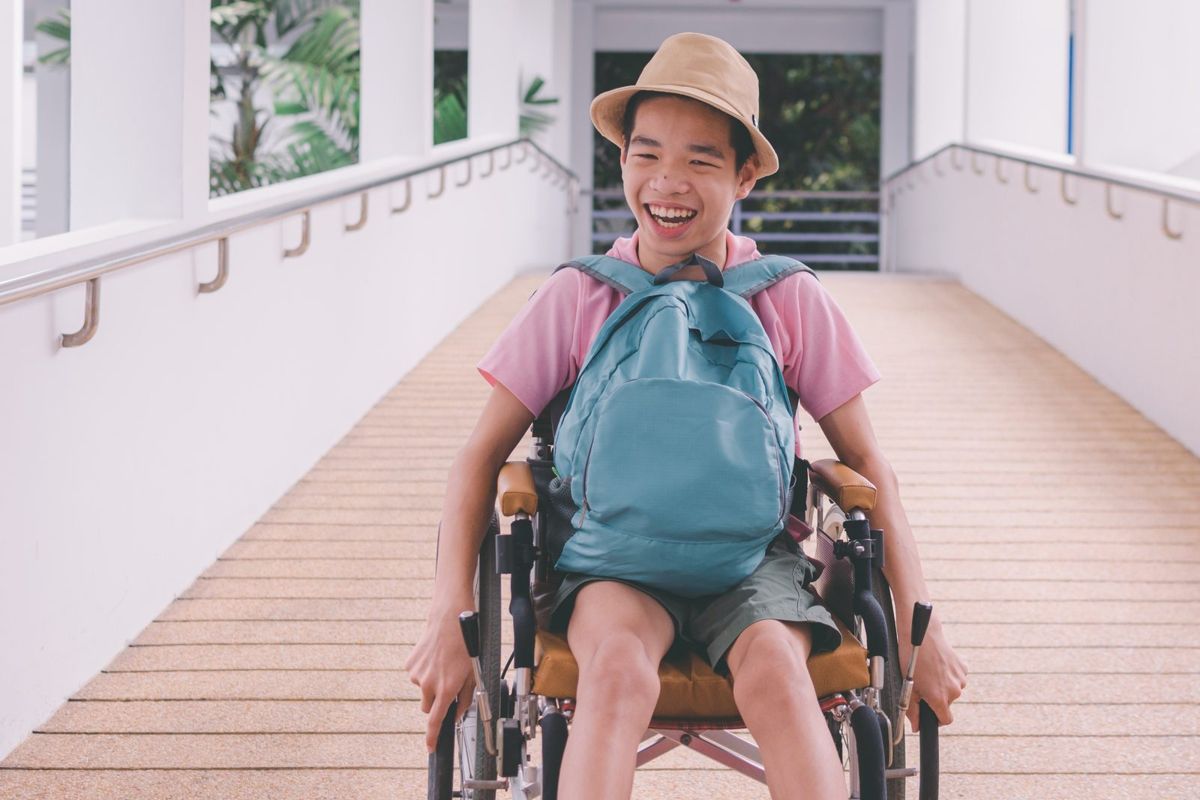 A smiling young man in a wheelchair wears a backpack.